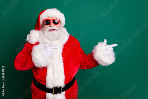 Photo of retired grandpa grey beard smile direct finger empty space offer newyear decorations wear red santa x-mas costume coat gloves sunglass belt headwear isolated green color background