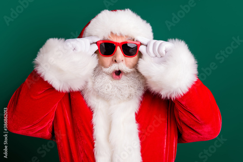 Closeup portrait photo of retired old man white beard open mouth hold eyewear shocked see unbelievable big sleigh wear red santa costume sunglass headwear isolated green color background photo