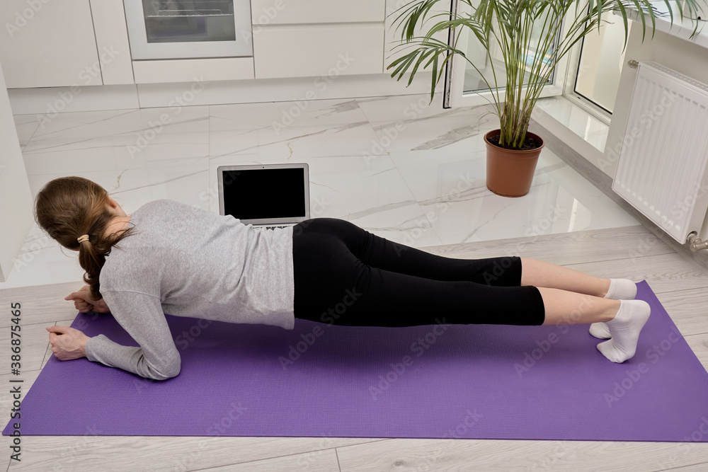 A blonde woman doing yoga stands in the plank position. Looks at the laptop screen. Training in sports and yoga online. There's a laptop on the floor. A blonde woman doing yoga stands in the plank position. Looks at the laptop screen. Training in sports and yoga online. There's a laptop on the floor.