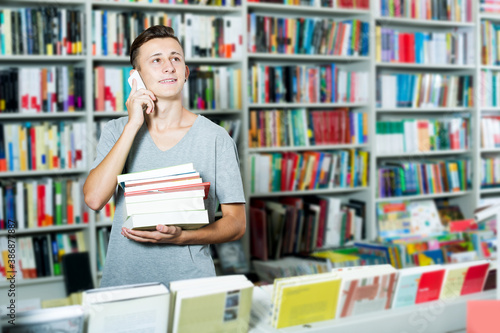 Cheerful teenage boy chatting on mobile phone and searching book in shop © JackF