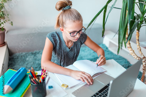 Beautiful young school girl left-handed working at home in her room with a laptop and class notes studying in a virtual class. Distance education and learning concept during quarantine
