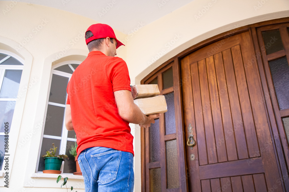 Caucasian deliveryman going to wooden door and holding boxes. Middle-aged male courier delivering order at home and carrying parcels in cardboard package. Express delivery service and post concept