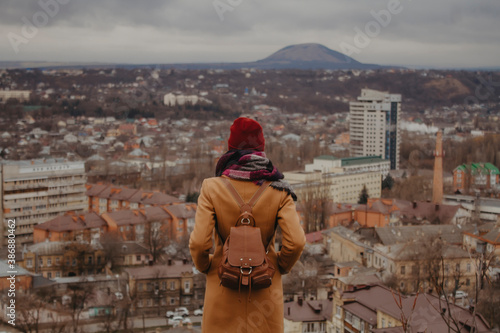 A woman in an autumn coat looks from the back at the old town and mountains.