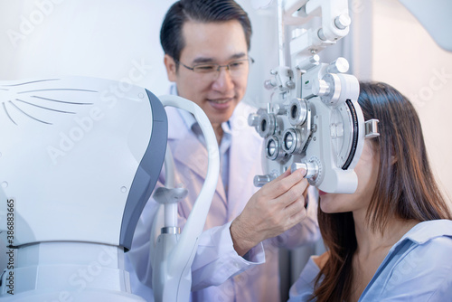 Asian woman looking through optical phoropter during eye exam, with an optometrist nearby, diagnostic ophthalmology equipment, selective focus
