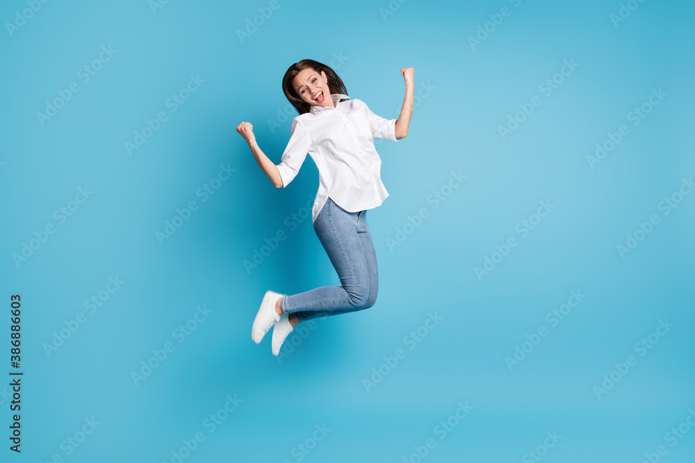 Full length photo of crazy lady jumping high up rejoicing raise fists wear white shirt jeans shoes isolated blue color background Full length photo of crazy lady jumping high up rejoicing raise fists wear white shirt jeans shoes isolated blue color background