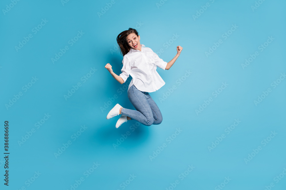 Full body photo of crazy lady jump high up raise fists victory wear white shirt jeans shoes isolated blue color background Full body photo of crazy lady jump high up raise fists victory wear white shirt jeans shoes isolated blue color background