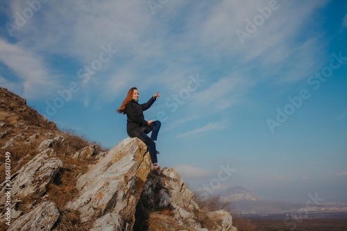 A young woman sits high on a mountain against a blue sky.