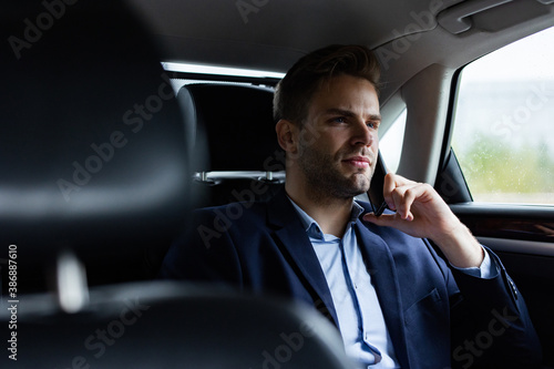 Close up portrait of young man in suit talking on the phone while sitting on the back seat of the car. Handsome young businessman test drive new car. © YURII MASLAK
