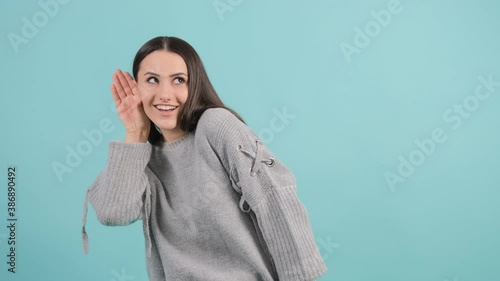 Close up of a curious young woman tries to hear you with her hand close to ear. The girl wants to hear the rumor and smiles, isolated over turquoise background.