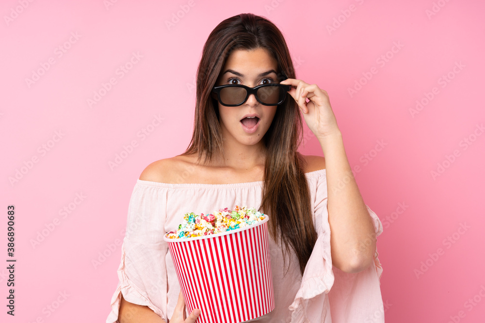 Obraz premium Teenager girl over isolated pink background surprised with 3d glasses and holding a big bucket of popcorns