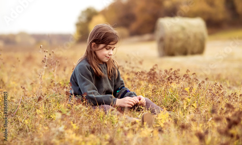 A teenage girl is sitting in a field on the grass against a background of hay.