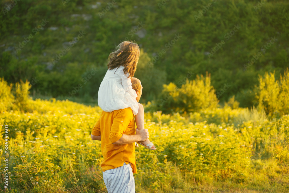 Father is carrying his daughter on shoulders walking on a field near forest in sunlight Father is carrying his daughter on shoulders walking on a field near forest in sunlight