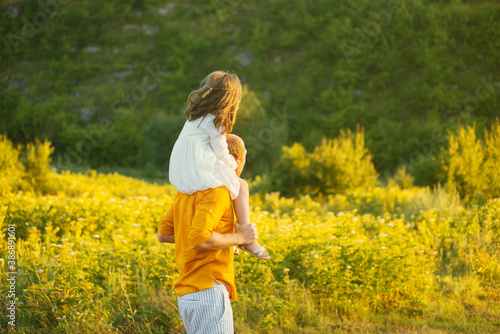 Father is carrying his daughter on shoulders walking on a field near forest in sunlight