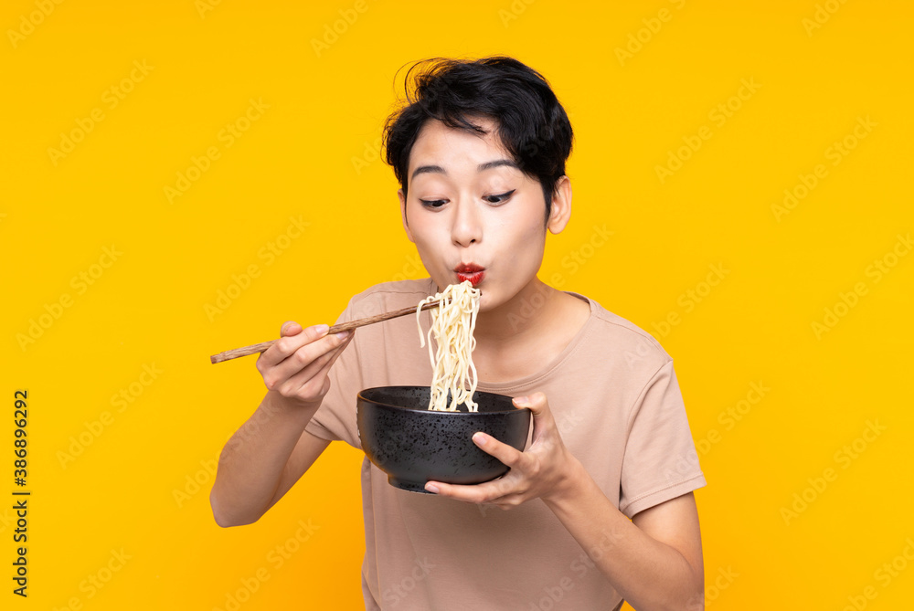 Young Asian girl over isolated yellow background holding a bowl of noodles with chopstick sand blowing it because they are hot Young Asian girl over isolated yellow background holding a bowl of noodles with chopstick sand blowing it because they are hot