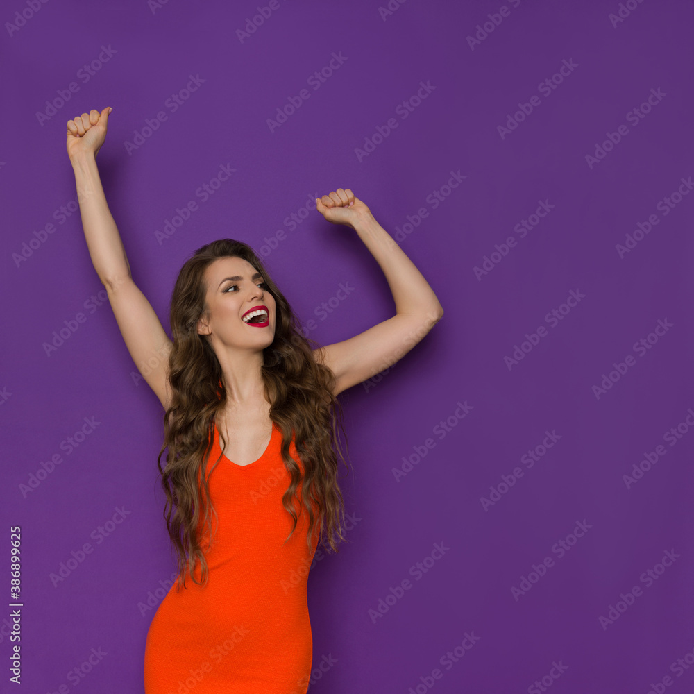 Carefree Young Woman In Orange Fitted Dress Is Holding Arms Raised And Laughing