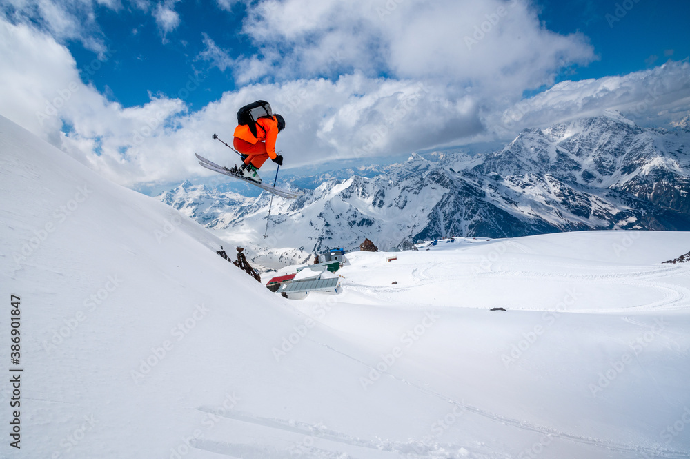 Professional athlete skier freerider in an orange suit with a backpack flies in the air after jumping on the lags on the background of blue snow and mountains
