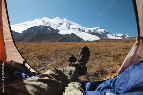 View of Mount Elbrus from an open tent with the legs of a tourist in mountain trekking boots