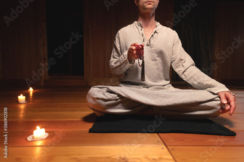 A man in gray linen clothes sits in lotus position in a dark meditation room and meditates with a red rosary in his hands. photo