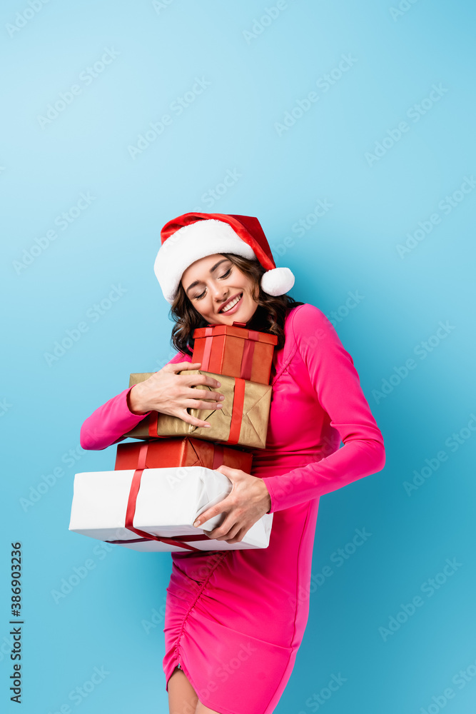 young joyful woman in santa hat and dress embracing wrapped presents on blue young joyful woman in santa hat and dress embracing wrapped presents on blue