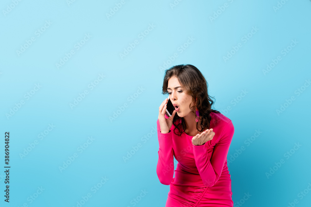 brunette woman in pink dress screaming while talking on smartphone and gesturing on blue