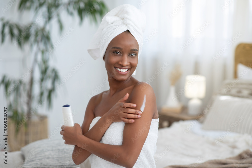 Focus on smiling young african american woman in towel, applies body moisturizer after shower Focus on smiling young african american woman in towel, applies body moisturizer after shower
