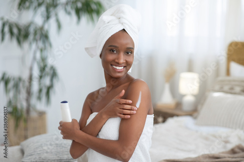 Focus on smiling young african american woman in towel, applies body moisturizer after shower