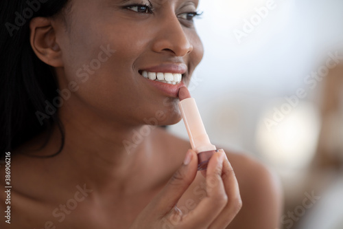 Focus on attractive millennial african american lady applying nude lipstick on lips and looks in mirror