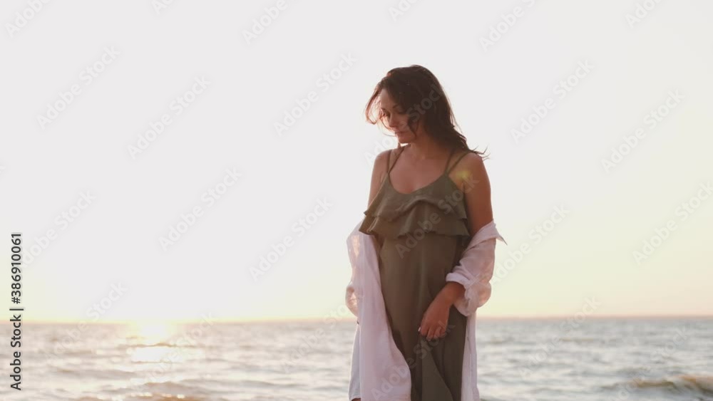 a young girl walks along the beach in a white shirt on a sunset background. Silhouette of a free girl in a white shirt walking along the seashore in the sunset a young girl walks along the beach in a white shirt on a sunset background. Silhouette of a free girl in a white shirt walking along the seashore in the sunset