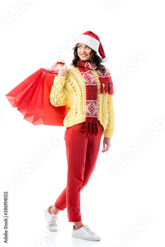 pleased woman in santa hat and scarf with ornament holding shopping bags isolated on white