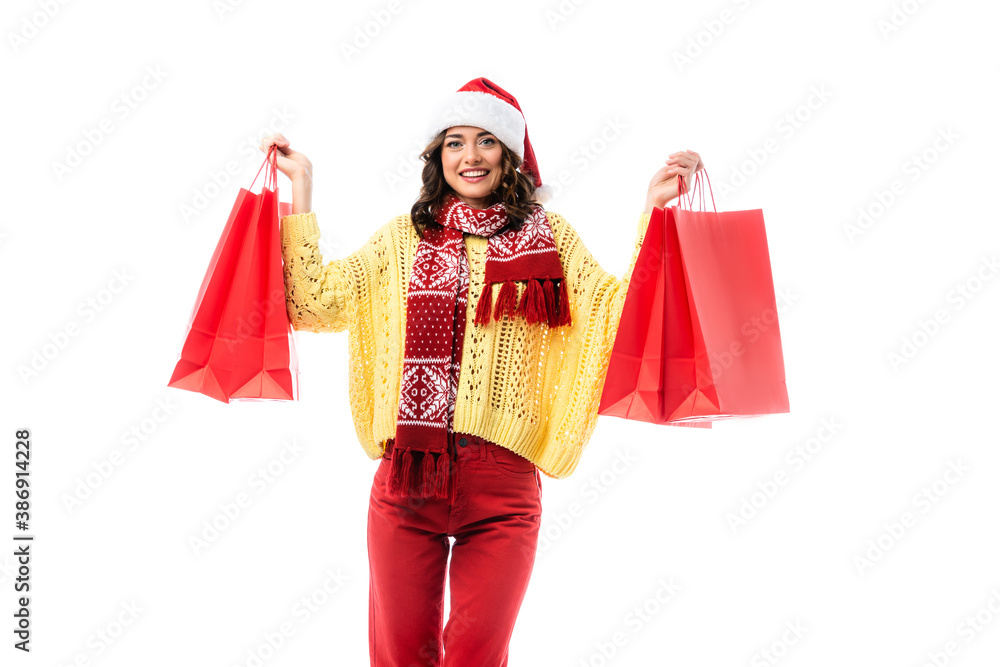 pleased woman in santa hat and scarf with ornament holding red shopping bags isolated on white pleased woman in santa hat and scarf with ornament holding red shopping bags isolated on white
