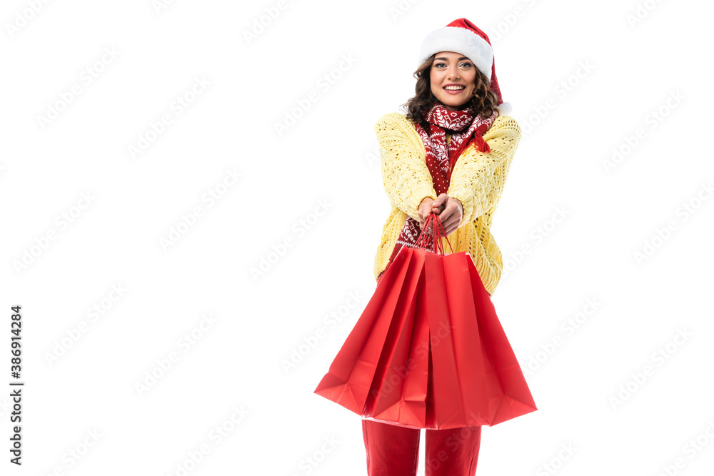 joyful young woman in santa hat and scarf with ornament holding red shopping bags isolated on white joyful young woman in santa hat and scarf with ornament holding red shopping bags isolated on white