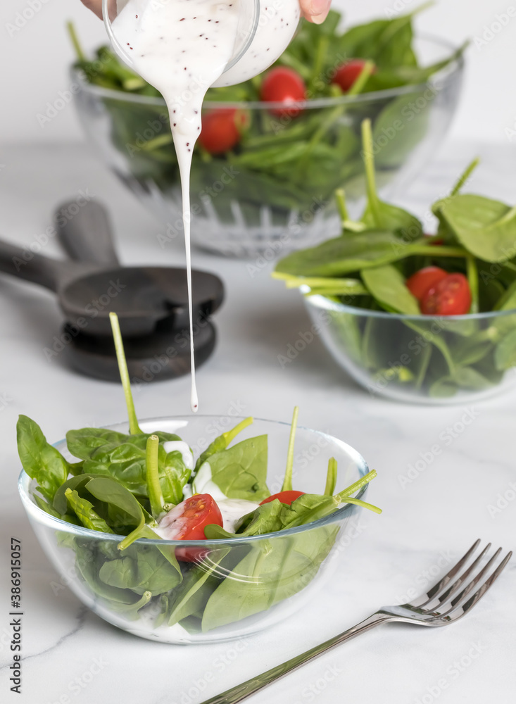 Close up of poppyseed dressing being poured into a spinach and tomato salad with other bowls of the salad in behind. Close up of poppyseed dressing being poured into a spinach and tomato salad with other bowls of the salad in behind.