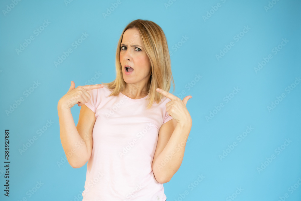 Fototapeta premium Portrait of young smiling woman in casual t-shirt pointing fingers herself isolated over blue background