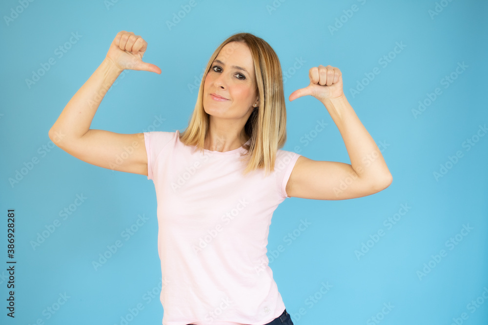 Fototapeta premium Portrait of young smiling woman in casual t-shirt pointing thumbs herself isolated over blue background
