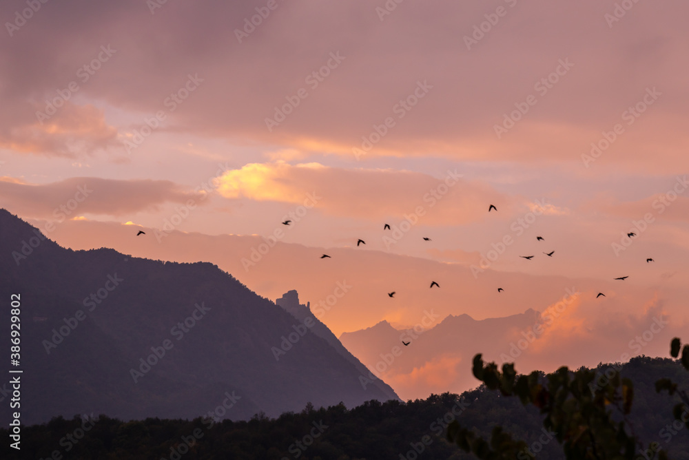 Avigliana, Italy. October 10th, 2020. View of Mount Pirchiriano at sunset. A flock of birds flies high in the sky with the Alps and the Abbey of San Michele in the background.