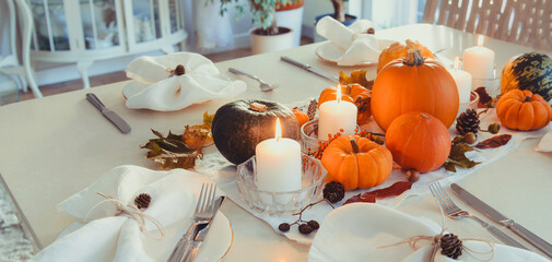 Festive table setting for Thanksgiving family home dinner. Fall composition with decorative pumpkins, nuts, cones, fallen leaves, and candles in light interior. Natural autumn decor. Selective focus