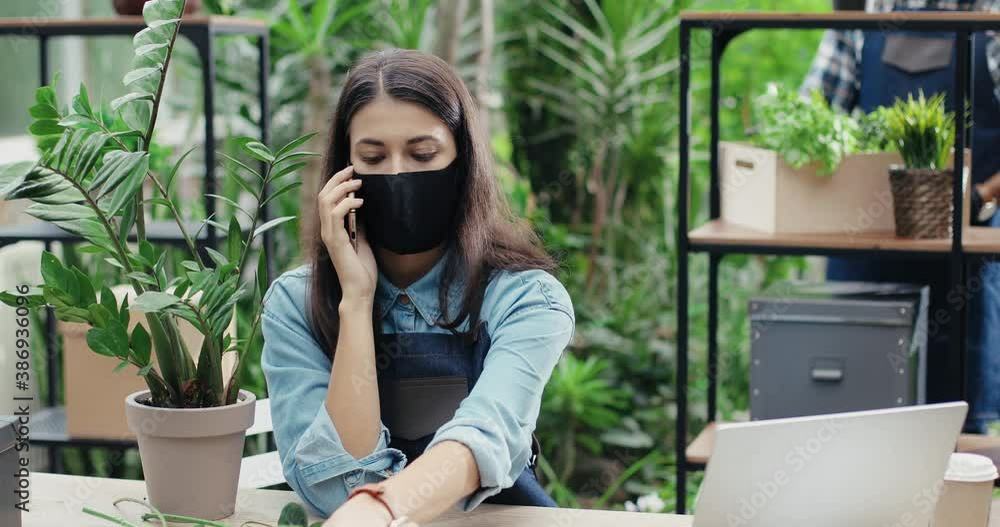Portrait of beautiful Caucasian woman worker in apron and mask talking smartphone and typing on laptop in flower shop. Female florist checking flowers indoors and calling on cellphone. Work concept Portrait of beautiful Caucasian woman worker in apron and mask talking smartphone and typing on laptop in flower shop. Female florist checking flowers indoors and calling on cellphone. Work concept
