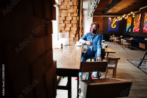 Stylish african american woman with dreadlocks afro hair, wear jeans jacket and face protect  mask at restaurant, hold cellphone. New normal life after coronavirus epidemic.