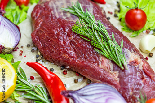 Raw red beef meat with green rosemary and fresh vegetables on light wooden cutting board background.