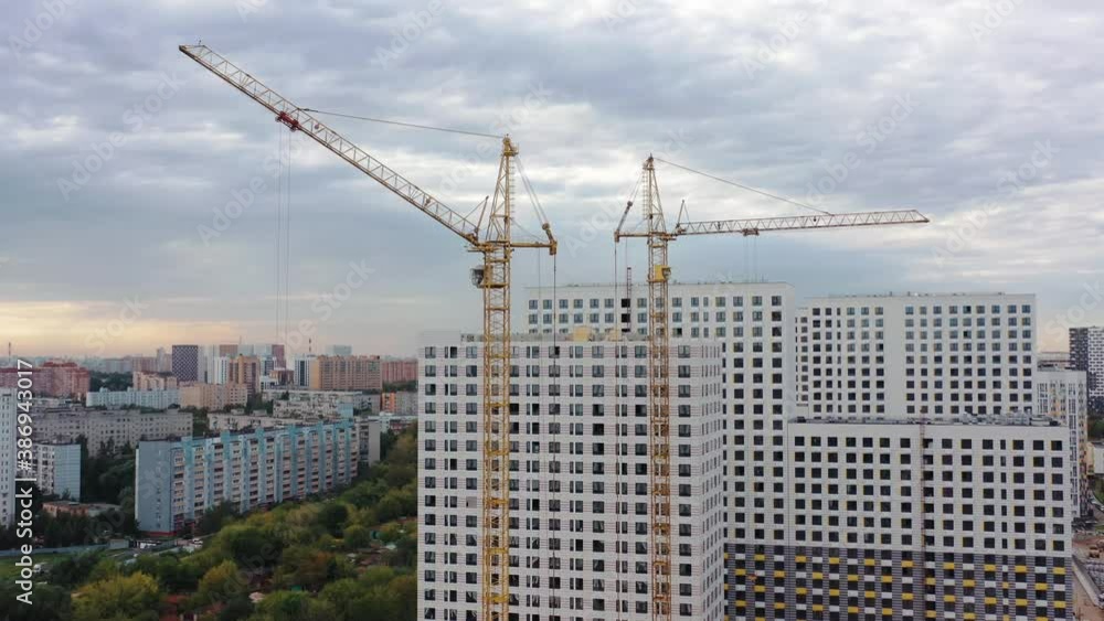 Buildings cranes and residential complex with high-rise apartment houses under construction, aerial drone tilt down shot. New apartment under construction, grey sky Buildings cranes and residential complex with high-rise apartment houses under construction, aerial drone tilt down shot. New apartment under construction, grey sky