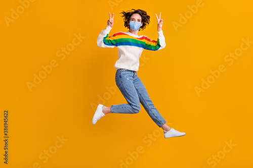 Full size photo of funny woman jump show v-sign wear mask sweater jeans footwear isolated on yellow background