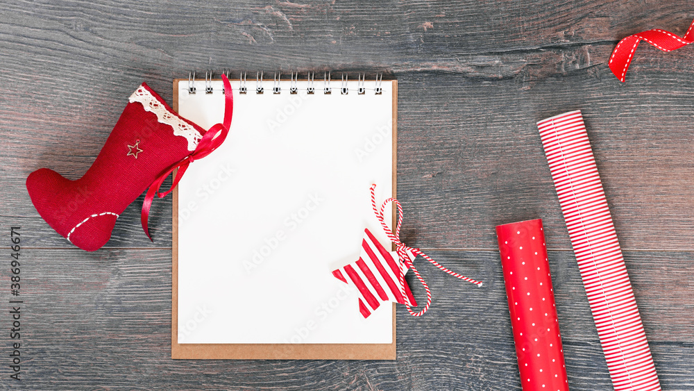 Fototapeta premium Merry Christmas mockup. Christmas stocking an open notepad on wooden background, red gift wrapping paper. New Year season greetings card resolutions mockup or goals. flat lay copy space