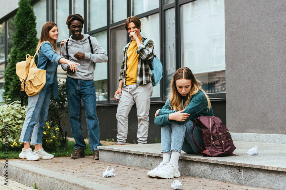 Fototapeta premium Selective focus of sad girl sitting near backpack, clumped paper and smiling multiethnic teenagers outdoors
