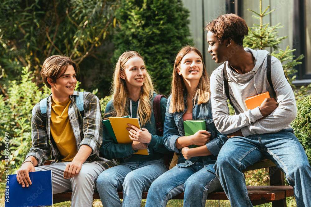 © LIGHTFIELD STUDIOS - Smiling teenagers with books looking at african american friend on bench outdoors