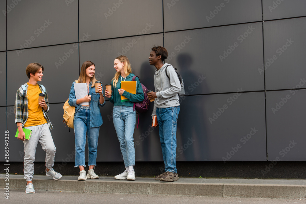 Fototapeta premium Positive multiethnic teenagers with notebooks and coffee to go talking near building outdoors