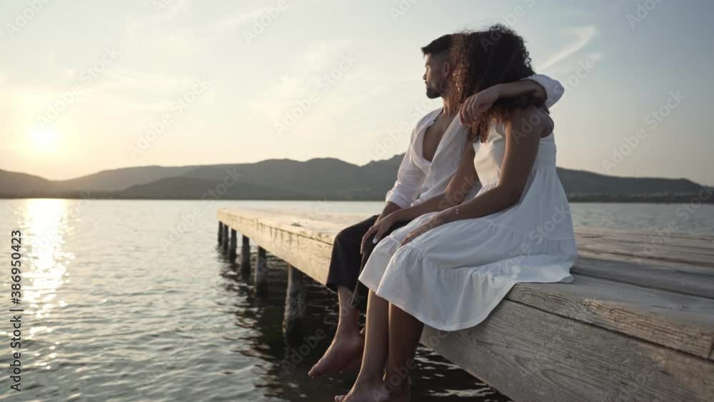 Romance scene of heterosexual mixed race couple looking at the setting sun sitting on a pier in the lake - Caucasian fine man embracing her black Hispanic curly girlfriend in a loving gesture Romance scene of heterosexual mixed race couple looking at the setting sun sitting on a pier in the lake - Caucasian fine man embracing her black Hispanic curly girlfriend in a loving gesture