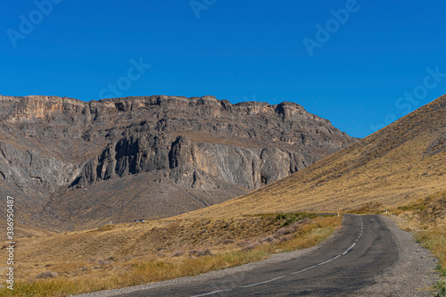 The road leads to the mountains. Mountain highway. Asphalt texture. Country highway. Mountains and hills in autumn. Mountain plants in autumn. Plants on the roadside. Dry yellow grass. Blue sky