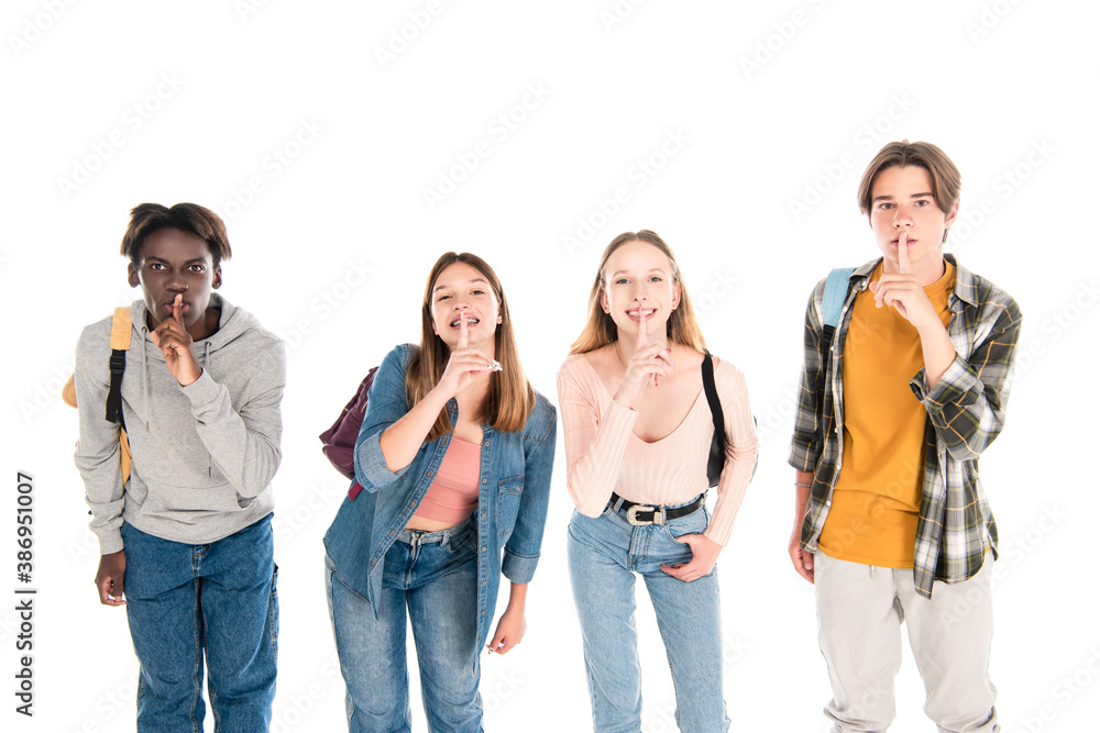 Obraz premium Smiling multiethnic teenagers with backpacks showing shh gesture isolated on white