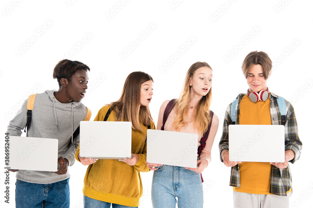 Excited multiethnic teenagers looking at laptop near friend with backpack isolated on white Excited multiethnic teenagers looking at laptop near friend with backpack isolated on white