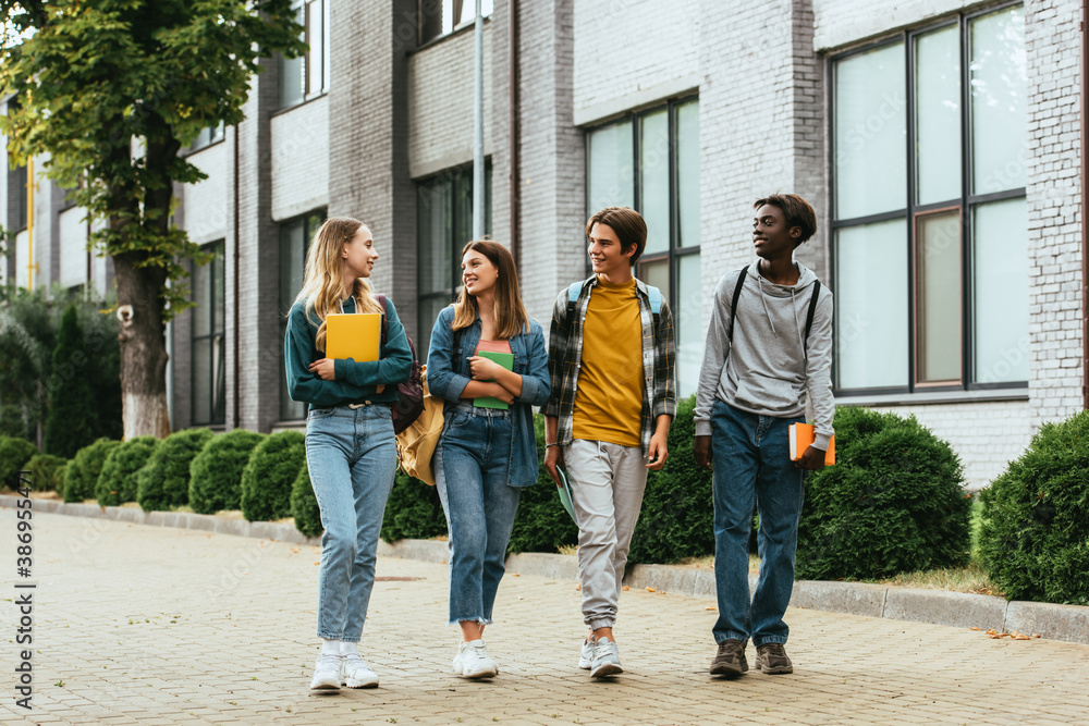 Fototapeta premium Smiling multicultural teenagers with notebooks and backpacks walking on urban street